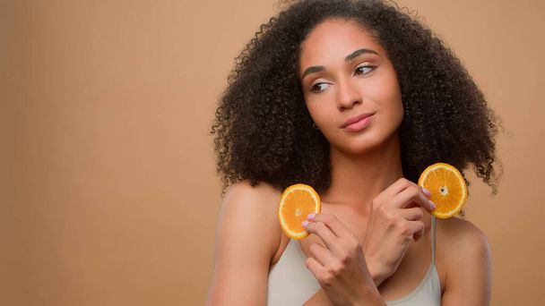 Hübsches ethnisches Modell Afroamerikanerin hält zwei Scheiben Zitrusfrüchte Frau hält die Hälfte der orangefarbenen Kosmetologie Hautpflege Haarpflege-Verfahren Diät Öko-Vitamin-Kosmetik ad beige Hintergrund - Foto, Bild