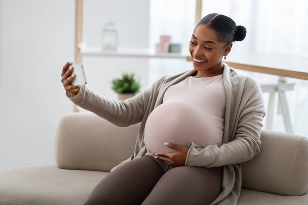 Wesoły Afryki młoda kobieta w ciąży siedzi na kanapie w domu, za pomocą smartfona, mieć spotkanie online z mężem lub przyjacielem, pokazując jej duży brzuch - Zdjęcie, obraz