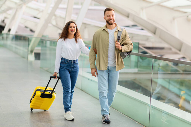 Расслабленная молодая пара, прогуливающаяся с багажом через терминал аэропорта, счастливые супруги тысячелетия, наслаждающиеся началом отпуска вместе, идущие к выходу на посадку, копировальная площадка - Фото, изображение
