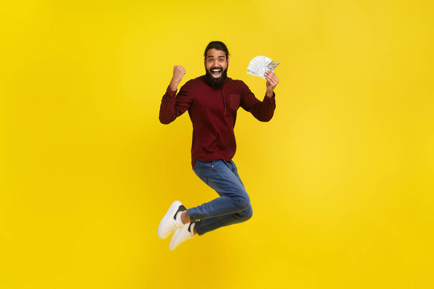 Happy indian guy winner jumping in the air with money in his hand, gesturing raising hand up, celebrating success, won lottery or got cashback, isolated on yellow studio background, full length - Foto, Imagem