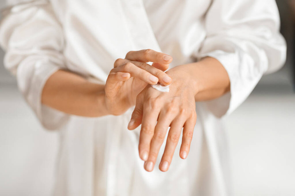 Selective focus on woman hands with nice manicure applying body cream. Cropped of lady wearing white silky bathrobe moisturizing her skin after shower, using hydrating lotion - Foto, immagini