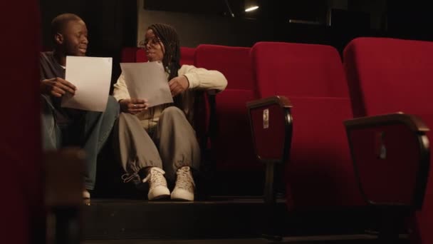 Low-Winkelaufnahme von jungen schwarzen Schauspielern oder Standup-Comedians, die auf Treppen im Theater mit roten Sitzen sitzen und plaudern, während sie den Monolog vor der Vorstellung überarbeiten. - Filmmaterial, Video