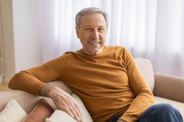 Удобный пожилой джентльмен, лежащий на диване, широко улыбающийся в комнате с легким, воздушным ощущением - Фото, изображение