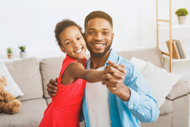 Pohledný otec a roztomilý dcera tančí doma, při pohledu na fotoaparát a s úsměvem - Fotografie, Obrázek