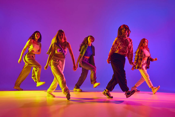 Imagem dinâmica de jovens mulheres talentosas em roupas casuais em movimento, dançando dança de rua contra fundo gradiente em néon. Conceito de juventude, dança de rua, dança contemporânea, moderna, dinâmica - Foto, Imagem