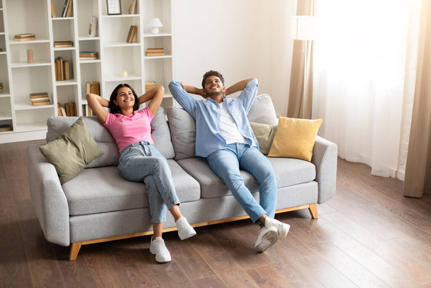 Unbeschwerte und glückliche indische Ehepartner, die es sich auf einem Sofa gemütlich machen, die Hände hinter dem Kopf, zufrieden und gemütlich in ihrem hellen, stilvollen Wohnraum - Foto, Bild