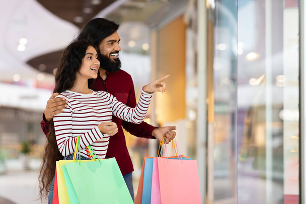 Romantický atraktivní tisíciletý indiánský pár ukazující na výlohu obchodu, procházky nákupním centrem, nošení barevných papírových tašek, užívání si prodejní sezóny, skvělé slevy, kopírovací prostor - Fotografie, Obrázek