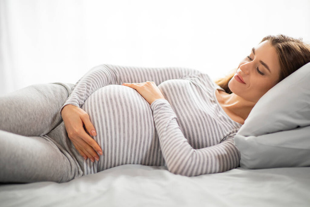 Mooie jonge zwangere vrouw omhelzen buik terwijl liggend op bed in de slaapkamer, glimlachende kalme moeder ontspannen thuis, genietend van gezonde zwangerschapstijd, close-up geschoten met vrije ruimte - Foto, afbeelding