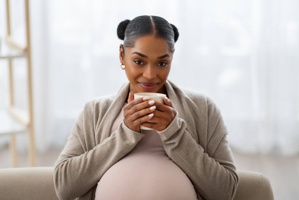Porträt einer fröhlich schönen jungen schwarzen Schwangeren, die es sich zu Hause auf dem Sofa gemütlich macht, Tee trinkt und in die Kamera lächelt. Fröhliches Schwangerschaftskonzept - Foto, Bild