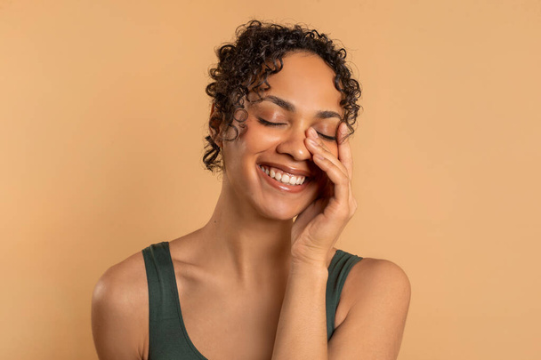Лечение кожи, косметики и лица. Счастливая латиноамериканка трогает лицо и улыбается, бежевый студийный фон - Фото, изображение