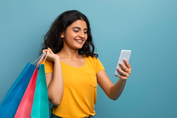 Błogosławiona młoda kobieta korzystająca z telefonu z torbami na zakupy w rękach, podkreślając cyfrowe zaangażowanie - Zdjęcie, obraz