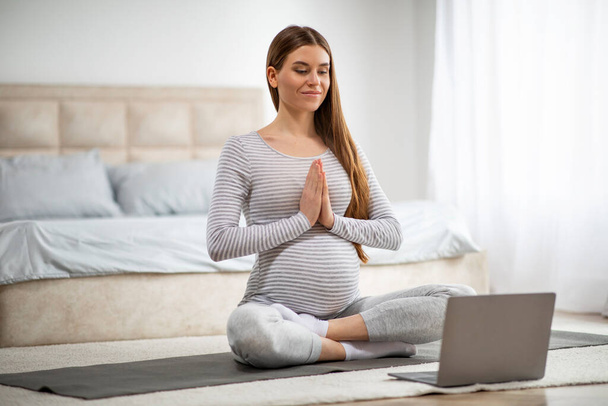 Спокойная молодая беременная женщина, занимающаяся йогой в Интернете, с ноутбуком дома, красивая будущая мать медитирует перед компьютером, сидит в позе лотоса на коврике и делает намасте Жест - Фото, изображение