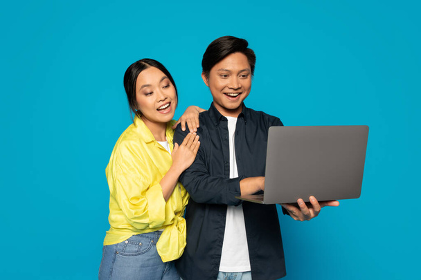 Überschwängliches asiatisches Millennial-Paar teilt sich einen Laptop-Bildschirm, die Frau stützt sich mit einem freudigen Lächeln auf die Schulter des Mannes und strahlt sowohl Glück als auch Zweisamkeit auf türkisfarbenem Hintergrund aus. - Foto, Bild