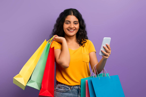 Жінка в жовтому верху посміхається тримаючи сумки для покупок і смартфон, пропонуючи позитивний досвід покупок - Фото, зображення