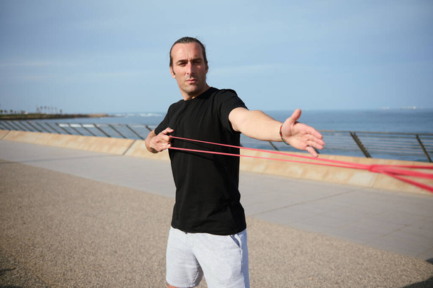 Young athlete doing arms, biceps and shoulder exercises with resistance fitness rubber bands on the beach outdoors. Handsome confident determined athletic man performs bodyweight training on promenade - Photo, Image
