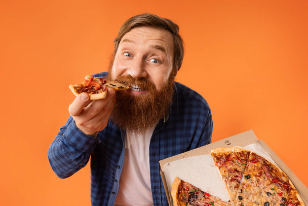 Brodaty rudowłosy facet Jedzenie plasterek smacznej pizzy pozowanie z pudełka Dostarczone z Pizzerii na tle Orange Studio. Portret jedzącego śmieciowe żarcie i zdradzającego posiłek. Odżywianie, przejadanie - Zdjęcie, obraz