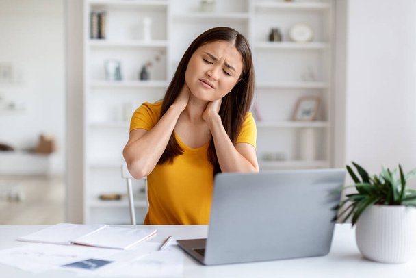 Mujer asiática estresada sintiendo dolor en el cuello, disgustada joven coreana mujer que sufre tensión mientras trabaja en el ordenador portátil, sentado en el escritorio con documentos en la oficina en casa, espacio para copiar - Foto, imagen
