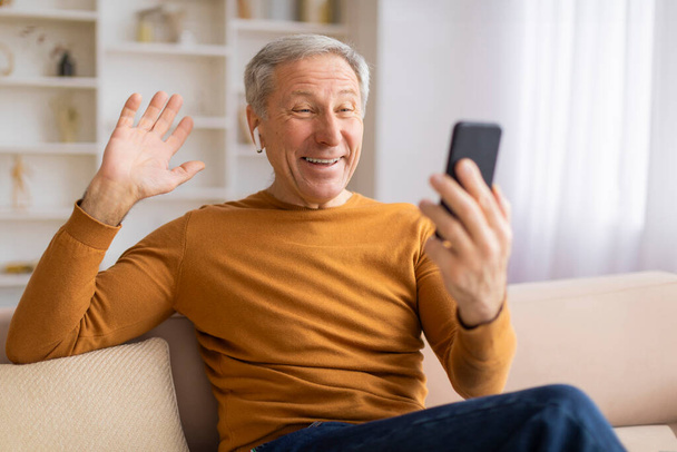 Счастливый пожилой мужчина, использующий смартфон для видеочата, с энтузиазмом машет на экран с радостным выражением - Фото, изображение