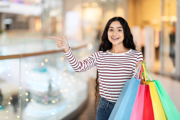Positive pretty long-haired young indian woman carrying colorful paper bags purchases, walking by modern city center, pointing at copy space. Black friday, season sale concept - Photo, Image