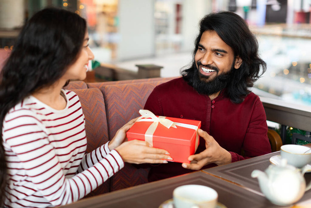 Mujer india joven de pelo bastante largo que presenta una caja de regalo envuelta a su novio emocionado en el café, haciendo una sorpresa especial. Amante pareja celebrando cumpleaños o aniversario en la cafetería - Foto, imagen