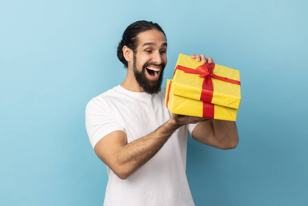 Portrait of extremely happy man with beard wearing white T-shirt looking into gift box, opening present and peeking inside with happiness. Indoor studio shot isolated on blue background. - Foto, Imagem