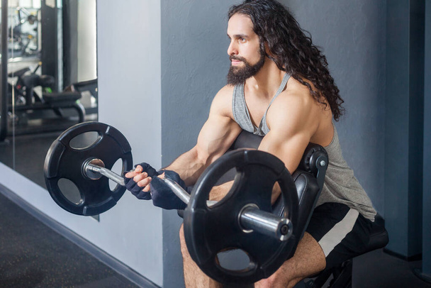 Portrait of muscular strong powerful man with long curly hair wearing sportswear training his body in gym lifting barbell, doing exercises for arms and shoulders. Indoor shot. - Fotó, kép