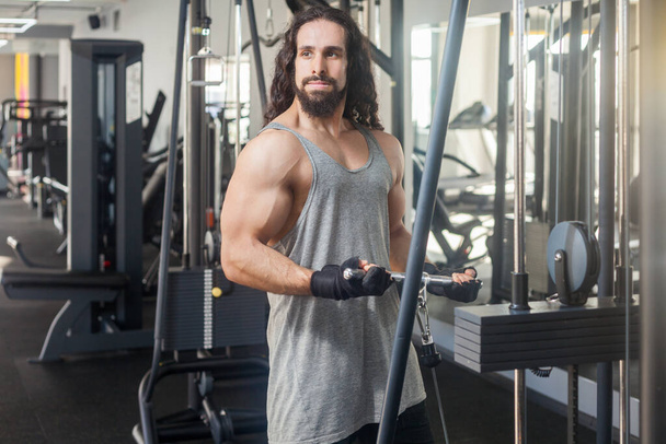 Portrait of muscular attractive man with long curly hair training at gym lifting weights using simulator, doing exercises for biceps and triceps, looking away has workout for arms. Indoor shot. - Foto, afbeelding