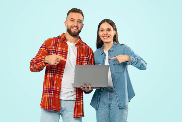 Glückliches kaukasisches Paar, das Laptop in der Hand hält und mit dem Finger auf den Computer zeigt, vor blauer Studiokulisse für freie Online-Arbeit und E-Learning-Angebote wirbt, in die Kamera lächelt - Foto, Bild