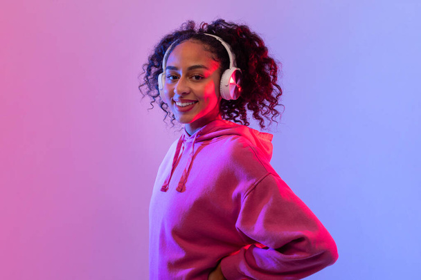 Smiling confident black woman with curly hair wearing white headphones and pink hoodie in vibrant neon pink and blue light, looking at camera - Photo, Image