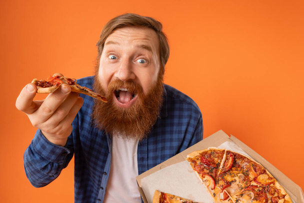 Hauska mies, jolla on punainen tukka ja parran syöminen Pizza Slice Standing, tilalla Pizzeria Box yli oranssi tausta studiossa, lähikuva muotokuva Shot. Nälkäinen kaveri nauttii roskaruokaa, huijata ateria - Valokuva, kuva