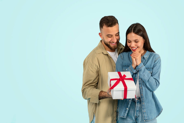 Happy young woman receiving wrapped gift box from loving husband, standing over blue studio backdrop and hugging. Millennial couple celebrating Valentines day or anniversary. Copy space - Photo, Image