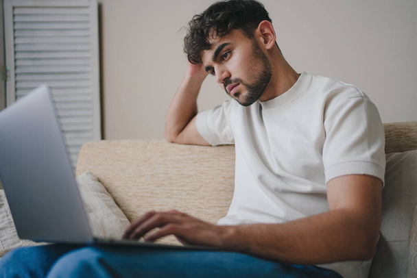Portret atrakcyjnego, młodego brodatego mężczyzny ubranego w luźne ubrania, siedzącego na kanapie w salonie, korzystającego z laptopa na czacie w sieci społecznościowej i - Zdjęcie, obraz