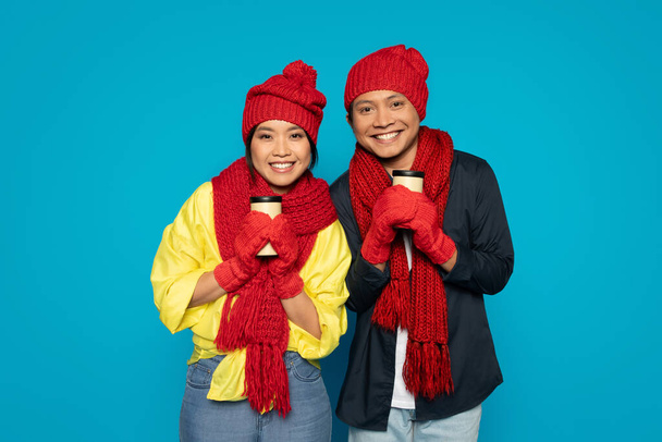 Cheerful funny millennial Asian couple in vibrant red winter hats and scarves smiling warmly while holding takeaway coffee cups against a striking turquoise background, studio - Photo, Image