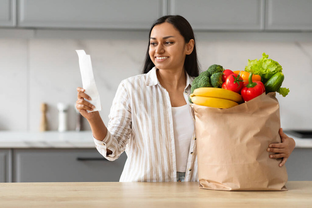 Jolie jolie jeune femme indienne avec sac en papier plein de fruits et légumes biologiques frais alimentaires debout à côté de la table de cuisine, chèque facture. Livraison d'épicerie, achats économiques - Photo, image