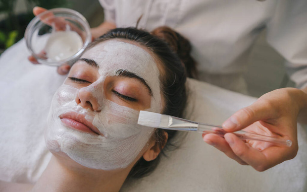 Kosmetikerinnen Hände mit Pinsel und Glasschale auftragen Schönheitsmaske auf das Gesicht der entspannten jungen Kundin. Kosmetisches Gesichtsbehandlungskonzept - Foto, Bild