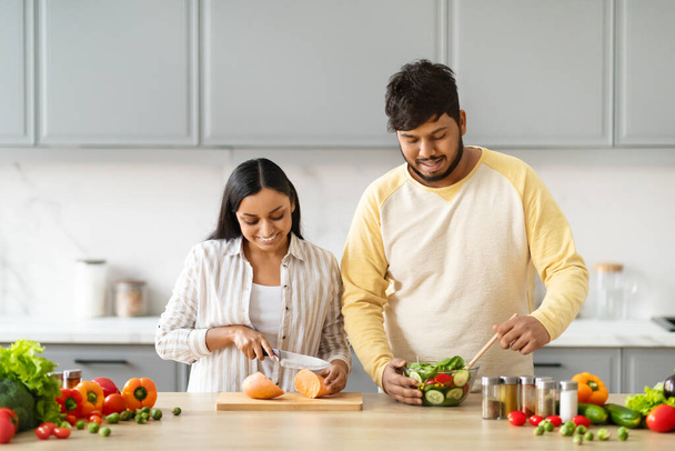 Joyeux couple indien cuisinant la nourriture et souriant préparer la salade pour le dîner ensemble dans la cuisine à la maison le week-end. Mari oriental et femme cuisinent le repas. Alimentation saine et recettes familiales Concept - Photo, image