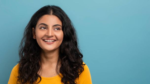 A joyful woman with long curly hair wearing a yellow t-shirt smiles gently against a solid blue background - Photo, Image
