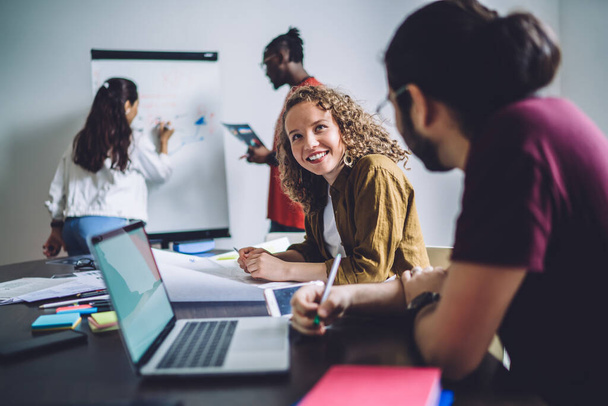 Low angle of carefree relaxed woman smiling away while talking with colleague sitting at laptop while dedicated multiracial coworkers analyzing work results writing on marker board in modern conference room - Foto, Imagem