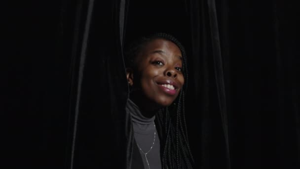 Pasie w górę radosnej młodej czarnej kobiety uśmiechającej się do machania przed kamerą, patrząc przez jedwabne czarne zasłony na scenie w teatrze - Materiał filmowy, wideo