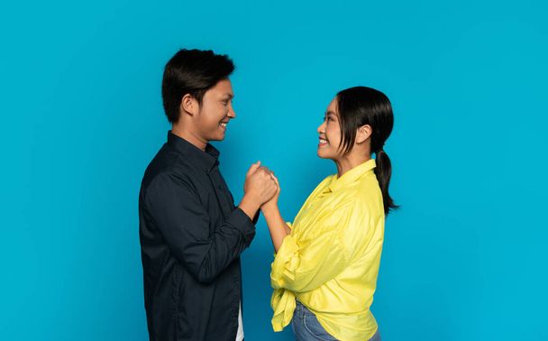 A joyful millennial japanese man in a dark shirt and a smiling woman in a yellow blouse hold hands, gazing at each other with a sense of happiness and connection against a blue background - Photo, Image