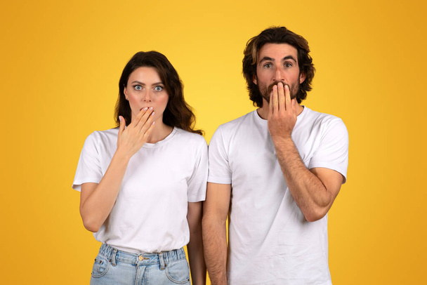Shocked millennial european man and a woman with surprised expressions, both covering their mouths with their hands, dressed in white shirts against a yellow background, studio - Photo, Image