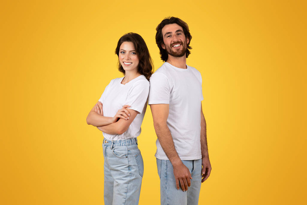 Egymás mellett, egy magabiztos európai Y-generációs nő és egy ragyogó férfi egymás mellett állnak, fehér pólóban, világos kék farmerban, napfényes sárga háttérrel, stúdióban. - Fotó, kép