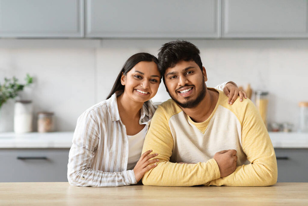 Retrato de amor jovem casal indiano posando juntos na cozinha. alegre milenar oriental mulher abraçando seu marido cônjuge namorado, sentado na mesa, sorrindo para a câmera - Foto, Imagem