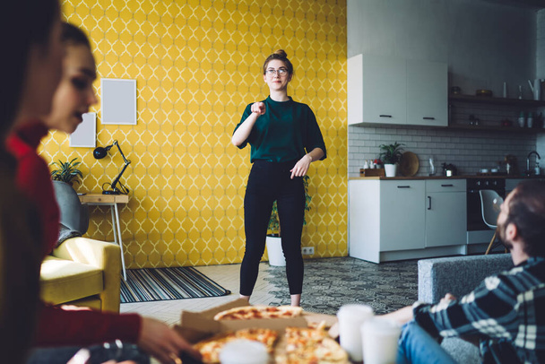 Playful female in casual wear thinking and pointing while playing game of charades with mates with pizza at modern home - Photo, Image