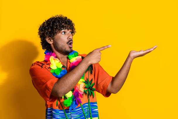 Portrait of speechless guy wear hawaii flower necklace look indicating at object on palm empty space isolated on yellow color background. - Fotografie, Obrázek