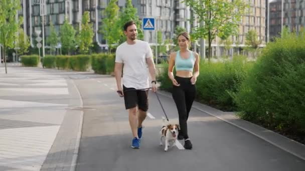 A positive sportive family spend leisure outdoors, walks and steps in unison with pet, a Jack Russell Terrier dog, on a warm summer day in the city park area. The concept of friendship and - Footage, Video