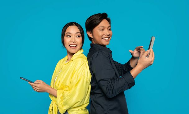 Dos individuos asiáticos milenarios espalda con espalda en ropa casual, hombre sonriendo mientras usa su teléfono, mujer con una expresión sorprendida también sosteniendo un teléfono, sobre un fondo turquesa brillante - Foto, Imagen