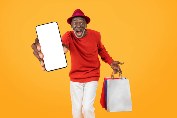 Экстатичный улыбающийся старший черный мужчина в красном свитере и шляпе, представляющий чистый экран смартфона и держащий красочные сумки для покупок, на оранжевом фоне, студия - Фото, изображение