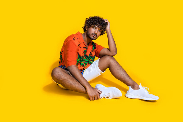 Full size photo of cool man dressed print shirt white shorts sitting on floor hand on head posing isolated on yellow color background. - 写真・画像