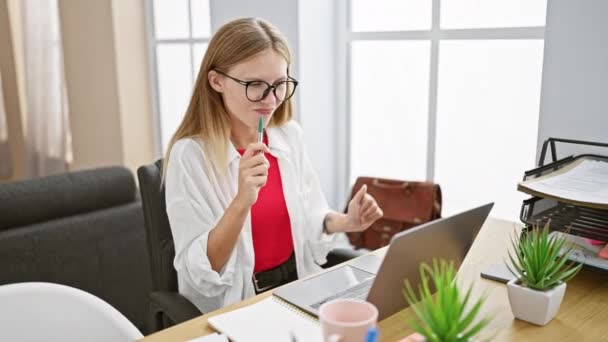 Задумчивая женщина в очках держит ручку в офисе с ноутбуком, растениями и оконным светом. - Кадры, видео